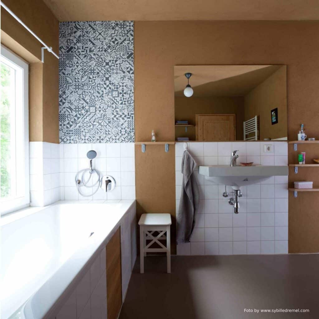 badezimmer mit teils weißen fließenflächen und lehmputz an wand und decke. großer spiegel über einem großen rechteckigen waschbecken. drei holzbrettchen übereinander als ablage. links großer wanne mit fließen und holzverkleidung. kleiner weißer hocker neben wanne. über der wanne längsseitig ein großes fenster mit grünem ausblick. brauner linoleumboden. im spiegel sieht man eine holzfüllungstüre in natur und einen weißen sprossenheizkörper.