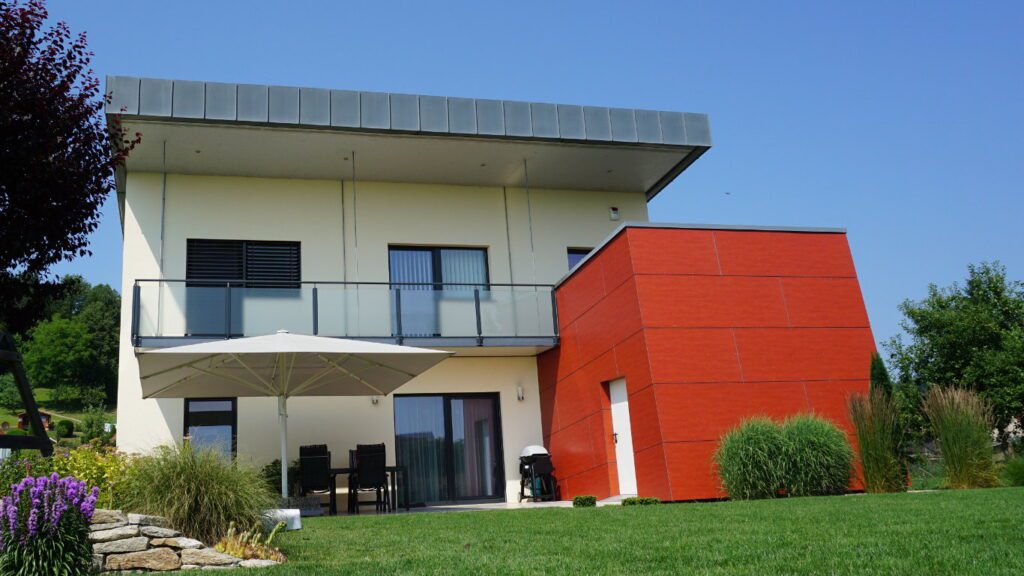zweigeschoßiges, hellgelbes pultdachhaus mit hoher seitlicher attikaverblendung aus vertikalen blechbahnen. balkon über die ganze front mit geländer mit milchiger glasbrüstung. rostroter kubus steht als nebentrakt rechts am hauptbaukörper. der kubus hat eine fassade aus plattenelementen und seitlich eine weiße türe von der terrasse aus, die duch diesen auf einer seite windgeschützt ist. ein großer weißer rechteckiger schirm steht vor der terrase. im vordergrund ein gepflegter rassen flankiert durch büsche,. gräßer, steinmäuerchen und mittelgroßen bäumen.