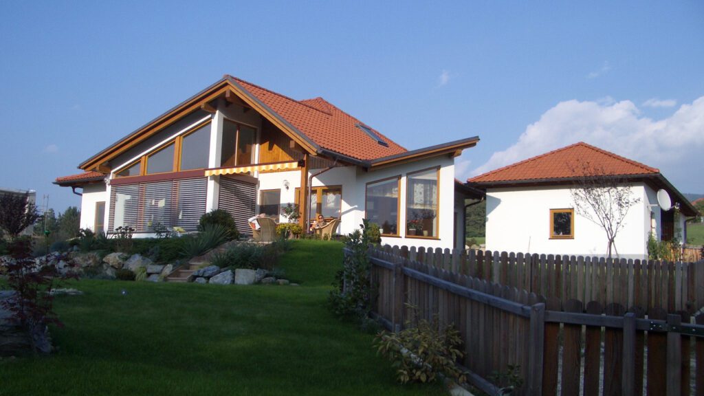 bungalow mit eckterrasse und dreieckigem wintergartenmit vogelartigem dach. zwei leute in korbstühlen auf der terrasse mit gelbweiß gestreifter markise. das gelände ist terrassiert. die abstufung ist mit schrägen zyklopenmauern gamacht. entlang des grundstücks ein holzbretterzaun. kanapp neben dem Bungalow mit weißer fassade und rotem ziegeldach, ein ebensolches kleines nebengebäude.