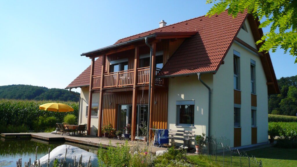 ein haus mit holz- und putzfassade steht direkt an einem teich. eine große glasschiebetür führt auf eine holzterrasse die leicht über den teich ragt. auf der terrasse stehen ein gelber schirm, ein tisch mit stühlen , ein wäscheständer mit blauem handtuch und topfpflanzen. im hintergrund ein hohes maisfeld und büsche. sommerlich trüber aber sonniger himmel. aufgeklapptes steildach mit roten dachziegeln. holzbalkon mit vier holzstützen die die überdachung über dem balkon und diesen selbst, tragen. sonniger himmel wolkenlos. gelber schirm auf der holzterrasse. niedriger metallzaun rechts am teich. großer weißer findling im vordergrund mit vielen zierpflanzen rundum. man sieht die blätter eines großen baumes von oben ins bild ragen.