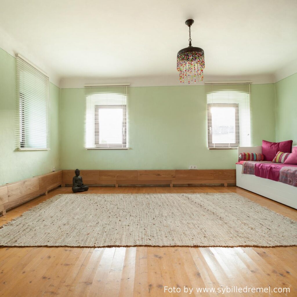 ein großer raum mit alten fenstern mit einem flachen boden oberhalb und innenliegenden geschlossenen jalousien. an der decke ein luster mit bunten glassteinen. rundum eine sockelleistenheizung rund 30 zentimeter hoch mit holzverkleidung. im eck hinten eine kleine sitzende buddhastatue. auf dem holzboden ein großer beigefarbener webteppich. rechts eine weiße küchenbank mit bunten pölstern und überwurf.