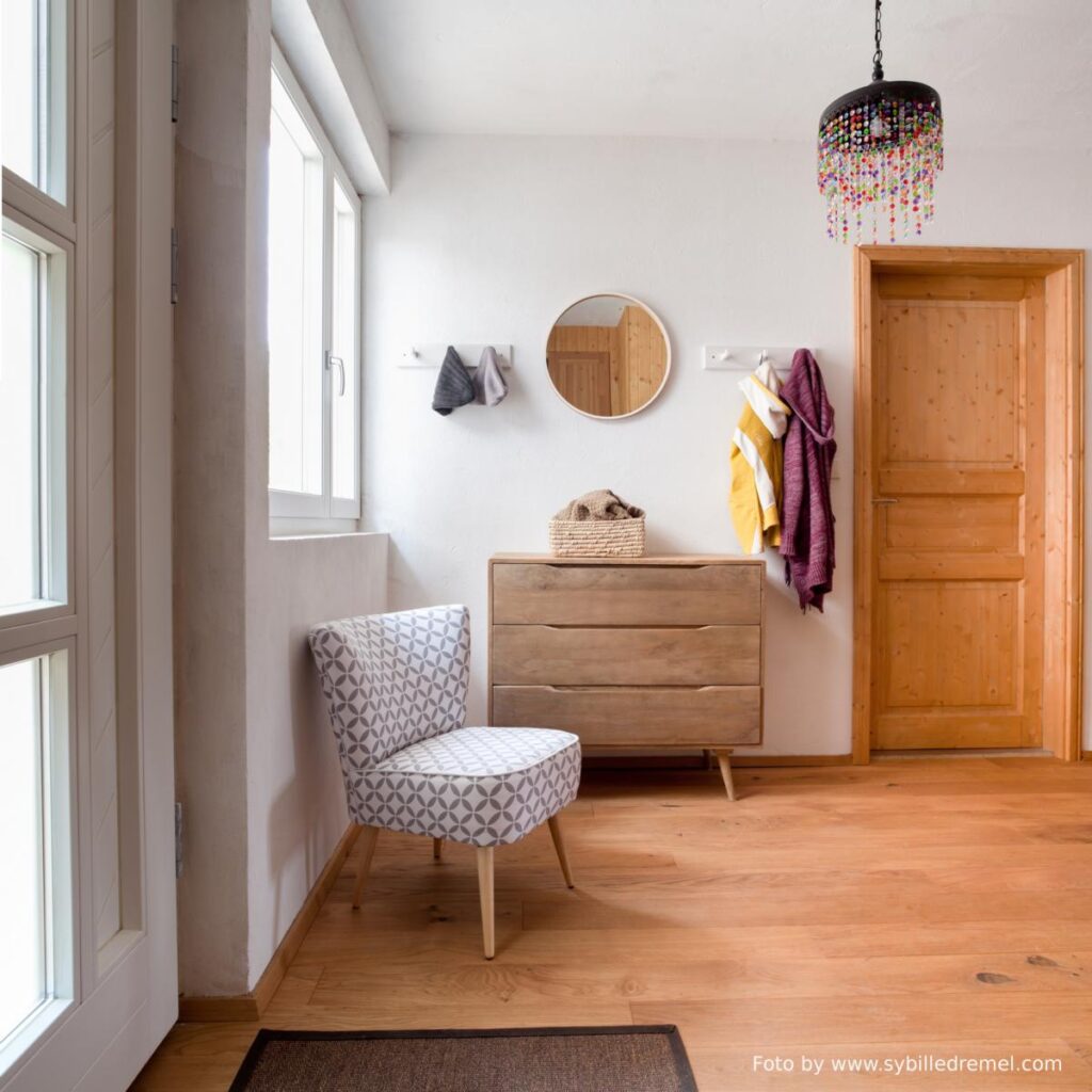 vorraum mit weißen putzwänden, holzboden, braunweiß gemusterten fauteuil, holzkommode mit drei langen schubladen, rundem spiegel, zwei garderoberelementen mit je drei haken mit zwei mützen und zwei hoodies drauf und glasluster mit bunten glassteinen. eine braune holzfüllungstüre in naturholz, eine eingangstüre mit weißem rahmen und glassegmenten und ein zweigeteiltes fenster mit weißem holzrahmen.