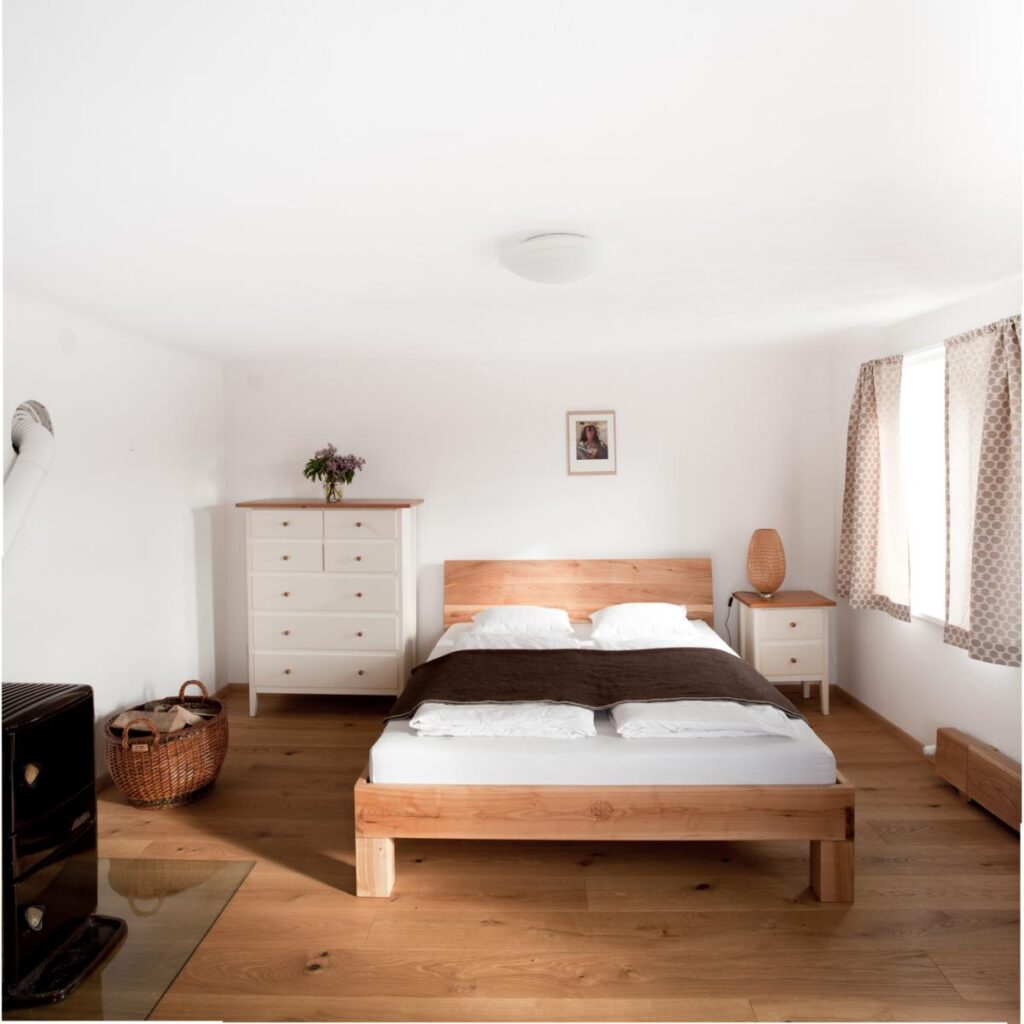 schlafzimmer mit weißen wänden, holzboden, braun gemusterten vorhängen, holzverschalter sockelleistenheizung, dunkelbraunen holzofen mit langem schrägen abzugsrohr, weiße, schubladenkasten und nachtkästchen. beide mit oberer holzplatte als abschluß. bett massiv aus holz mit rückenteil, weiß bezogenen mit brauner tagesdecke. nachtkästchenlampe mit geflochtenem schirm. kleines bild von einem indianer über dem betthaupt. großer korb mit zwei griffen mit brennholz gefüllt. milchige glaslampenschale mittig über dem bett.