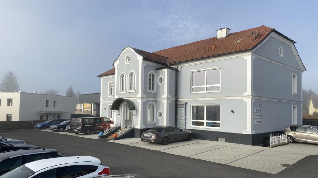 historistische zweigeschoßige villa mit grauer fassade und weißen putzfaschen. ein vorspringendes zweigeschoßiges eingangsportal in das eine freitreppe über 8 stufen führt. rechts am gebäudeeck ein weißes geschmiedetes geländer an einem kellerabgang. teilweise runde kleine fenster und ein geschwungenes rundes dach über dem eingangsbau. en großer parkplatz mit asphalt im fahrbereich und grauem pflaster bei der parkierung. links hinten ein einfaches weißes haus mit weißer fassade. blauer himmel. leicht nebelig.