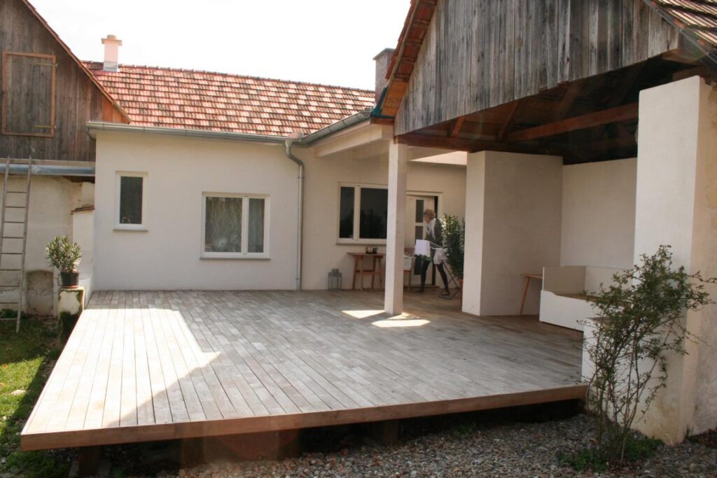 großé leere holzterrrasse mit blick auf ein L-förmiges bauernhaus. über eine leiter geht es zu einer holztüre im dachspitz. rechts eine mauernische mit küchenbank. ein holzdach ragt über die mauernische. vorne ein streifen von einer schotterfläche. im hintergrund in einem durchgang behängt eine frau einen wäscheständer mit weißer wäsche. über ihr ein flachdachelement mit oberlichtfenster.