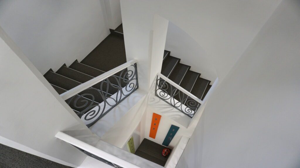 viereckkiges stiegenhaus mit grauem teppichbelag, gemauerten innenpfeilern und eckpodesten. blick von oben ins rund ein mal ein meter großes stiegenauge. flache bögen über dem innenliegenden geländer strecken sich von ecksäule zu ecksäule. ganz unten im stiegenauge sieht man bunte vertikale bretter mit garderobenhaken. ein feuerlösche steht auch ganz unten in dieser garderobennische. die stiegengeländer sind mit spiralförmigen stahlstangen gefüllt. der handlauf ist aus lackiertem holz. die wände sind weiß.