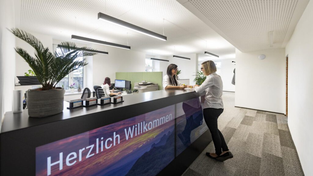 lange theke als empfang in einem bürogebäude. zwei frauen sprechen miteinander. herzlich willkommen auf der front der theke. im hintergrund ein großraumbüro.