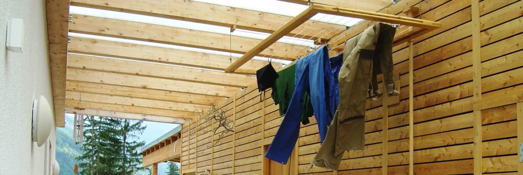 gang zwischen einem gebäude und einem nebengebäude mit horizontaler lammellenschalung aus holzlatten. der gang ist mir einem glasdach überdacht, man sieht viele holzsparren für das glasdach. von den holzbalken ist ein trockenrahmen mit wäscheleinen abgehängt. auf diesem sind arbeitshosen und tshirts. hinten ein bewaltetes tal mit nadelbäumen. links zwei runde wandleuchten an der weißen putzfassade des hauses.