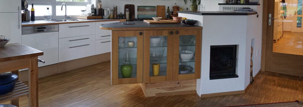 weiße küche mit auskragendem holzkächstchen mit verglaster dreiteiliger front mit schüsseln als inhalt. von außen zu beheizender kachelofen in die küchenzeile integriert. der hauptteil der küche ist mit lackierten weißen fronten und einer holzarbeitsplatte. ein geteites fenster mit fixverglasung unten im hintergrund. rechts hinten geht eine glastüre in den gang mit weißen schuhschränken und gelblicher beleuchtung.