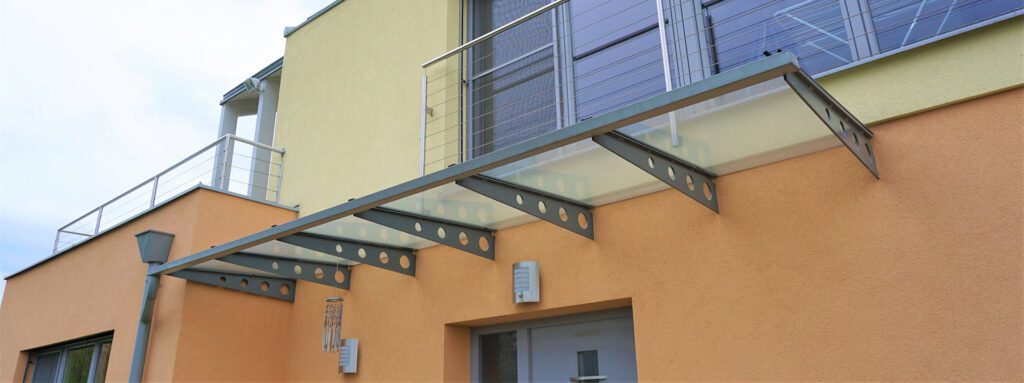 gelbe und lachsfarbene putzfassade. unten sieht man noch etwas von der grauen eingangstüre. über dieser die eingangsüberdachung aus verzinkten stahlschwertern mit kreisrunden bohrungen und einer glasabdeckung. im Obergeschoß ein mehrteiliges großes, raumhohes fenster mit stahlgeländer mit seilverspannungen. links oben eine terrasse mit ebensolchen geländer. ein windspiel am vordach.