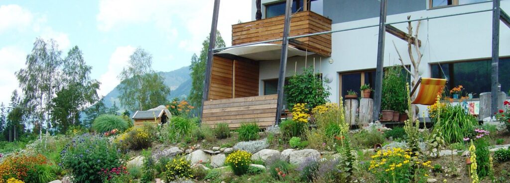 einsteingarten mit vielen verschiedenen blühenden pflanzen steigt zu einer hausterrasse auf. das haus hat einen kleinen holzbalkon und vier hohe stützen davor, die das weit auskragende dach tragen. ain gelboranger hängestuhl ist auf den stützenverstrebungen angebracht. ein kleines weißes sonnensegel schützt den sitzbereich mit seitlicher windschutzwand und vorderseitigem geländer zu, steingarten hin.