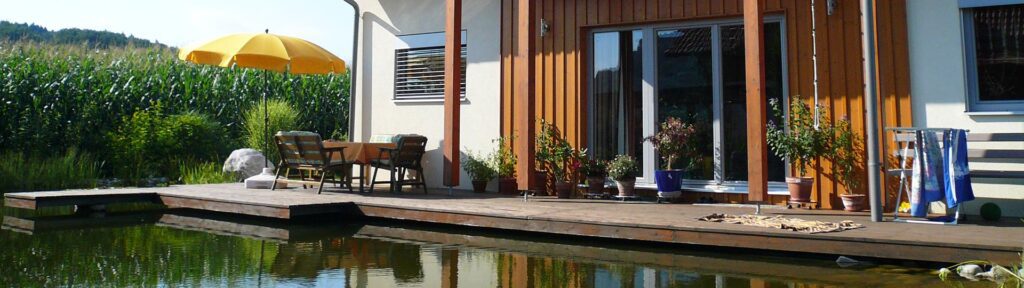 ein haus mit holz- und putzfassade steht direkt an einem teich. eine große glasschiebetür führt auf eine holzterrasse die leicht über den teich ragt. auf der terrasse stehen ein gelber schirm, ein tisch mit stühlen , ein wäscheständer mit baluem handtuch und topfpflanzen. im hintergrund ein hohes maisfeld und büsche. sommerlich trüber aber sonniger himmel.