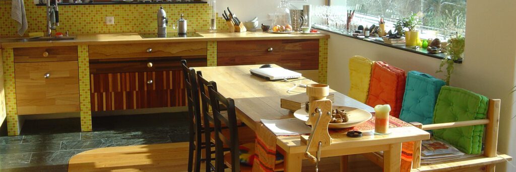 eine selfmadeküche mit arbeitsplatte aus holz. zwischen den küchenunterkästen sind gemauerte teiler mit gelben fließen an der front. ein langer holztisch mit holzbank und sesseln mit bunten pölstern. eine getreidemühle ist am tisch angeschraubt. ein laptop und eine kerze sind am tisch. ein fenster bietet ausblick ins grüne, am herd stehen kaffeemaschienen. der boden ist mit schwarzen steinplatten und holzbrettern belegt.