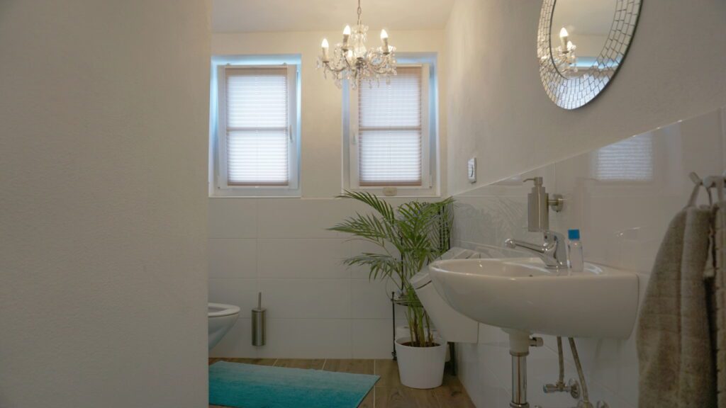 toillette mit zwei kleinen fenstern. an der decke ist ein luster mit 5 lampen. im vordergrund ein waschbecken. im eck steht ein zimmerfarn in einem topf.